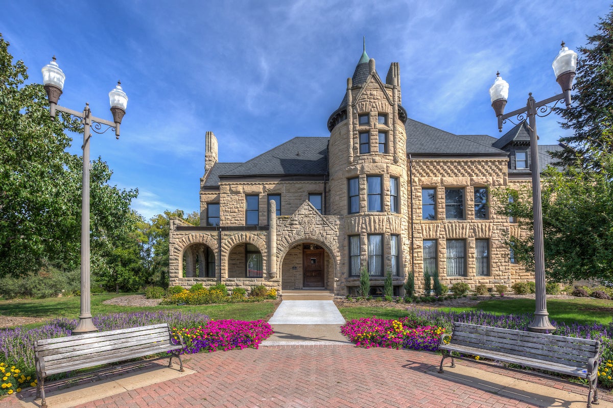 Front view of the Louis Smith Tainter House, a historic tan stone building with arched entryways and a turret, framed by flower beds, benches and decorative lampposts under a blue sky.