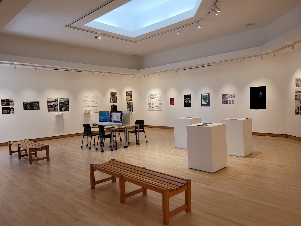 A gallery with various artworks, some digital media and games, podiums with a few books, as well as two tv's on the wall.