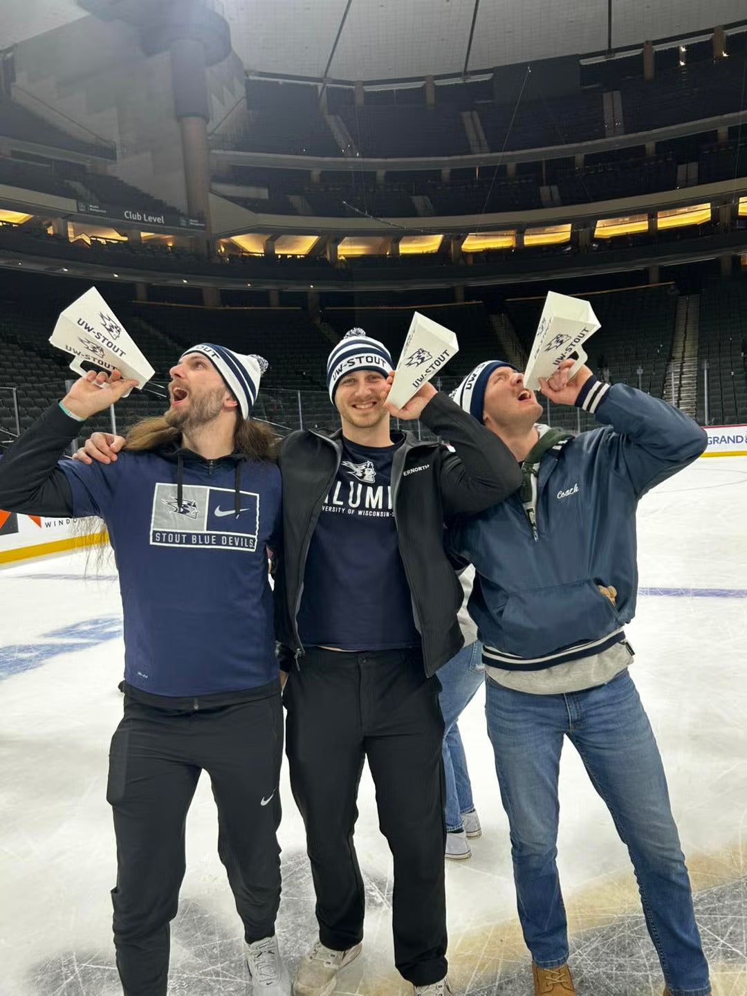 Three people stand on an indoor ice rink, each holding a white UW‑Stout megaphone near their mouth. All three wear UW‑Stout winter hats and dark blue or black UW‑Stout apparel, including one shirt that reads “Alumni” and another that reads “Stout Blue Devils.” Stadium seating rises in the background under bright arena lighting.