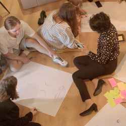 Group of people kneeling on the floor, collaborating and sketching ideas on a large sheet of paper with sticky notes nearby.