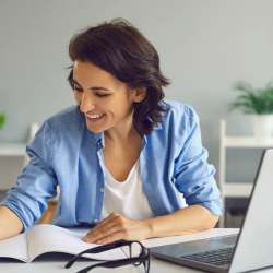 A woman looking down at a book with an open laptop