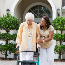 A younger woman smiling and assisting an older woman using a walker as they walk together outside a building entrance.