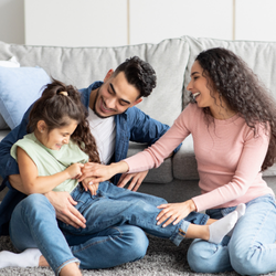 A mother, father, and their young daughter sit together on a gray living room rug in front of a light gray sofa. The family is casually dressed and laughing as the parents playfully interact with the giggling child, who is sitting between the father's legs