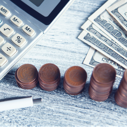 A close-up view of a silver calculator next to five stacks of copper coins that get taller from left to right. A white pen sits below the coins, and the corners of several one-hundred dollar bills are visible in the top right. All of the items are resting on a weathered white wooden table.