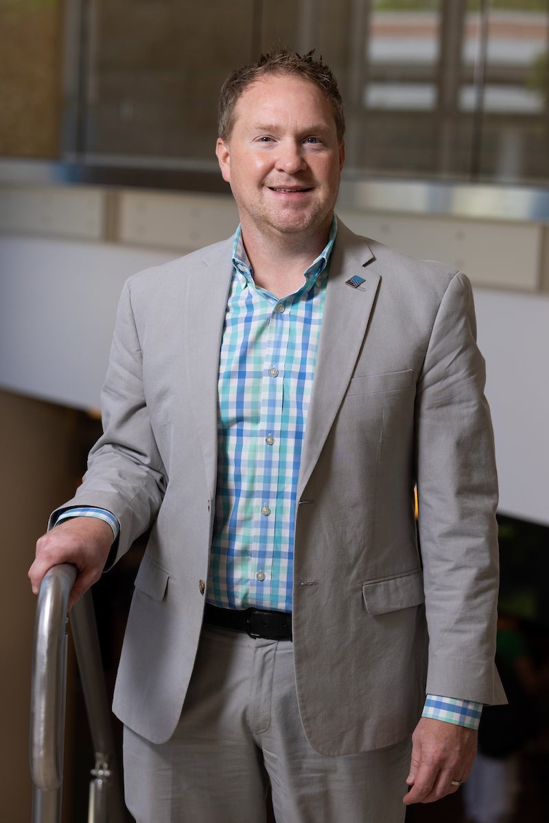 Person wearing a light gray suit jacket and checked button-down shirt, standing indoors with one hand resting on a railing.