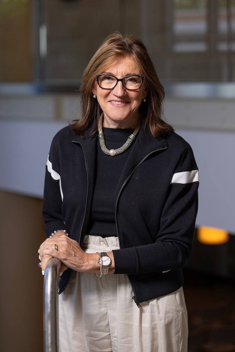 Person wearing a black zip-up jacket with white stripes, a black top, and light-colored pants, standing indoors with hands resting on a railing.