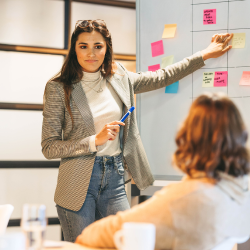A woman pointing at a sticky note on the borad, with a blue marker on her other hand