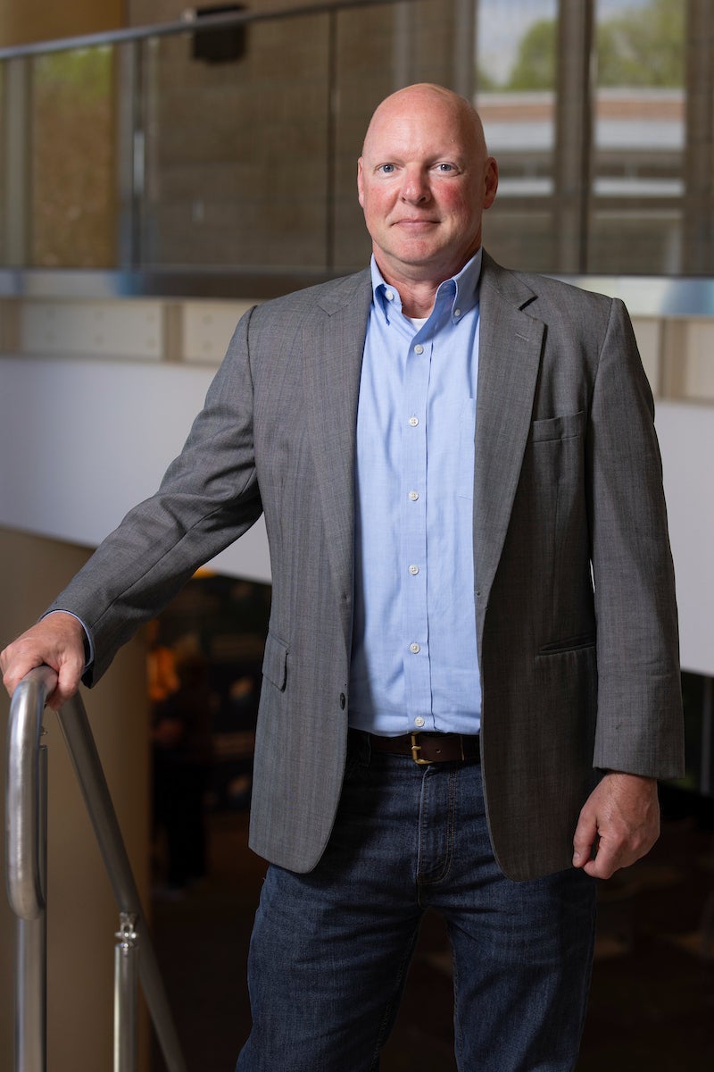Person wearing a gray blazer, light blue button-down shirt, and jeans, standing indoors and resting one hand on a railing.