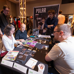 Attendees at an event gathered around a black table playing a tabletop game. The table is covered with game tiles, cards, and small tokens, and a large poster behind them reads Polar Void. A woman in a white shirt and a man in a black shirt are pointing at the board layout, while another man stands behind them with his arms crossed observing the gameplay. Small white signs with the letters SGX are visible on the front edge of the table.
