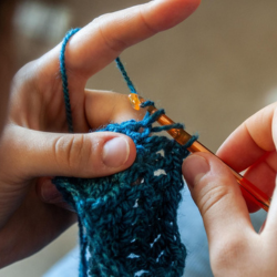 Close up of two hands crocheting a piece of dark blue yarn with an orange crochet hook.