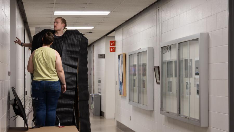 One student standing on a latter faces her groupmate, who stands over a foot taller while he is using stilts. He's in long black panels of materials that flow around him as he walks.