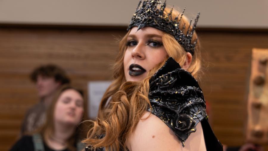 Female student in her project outfit turns and looks over her shoulder at the camera. She has a black spikey crown, with spike black pieces covering her shoulders.