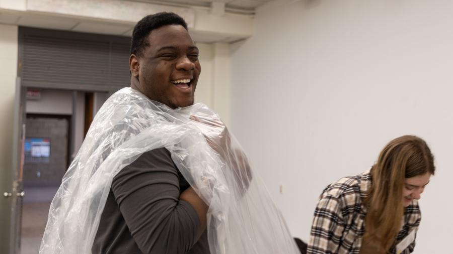 A student looks at the camera as he laughs and puts his head through a large pastic sheet so it would lay on him like a shirt.