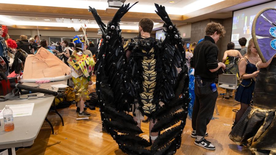 A student stands in the green room and on his back sit large, covered in black feathers and googley eyes, mechanical wings that are taller than the student.