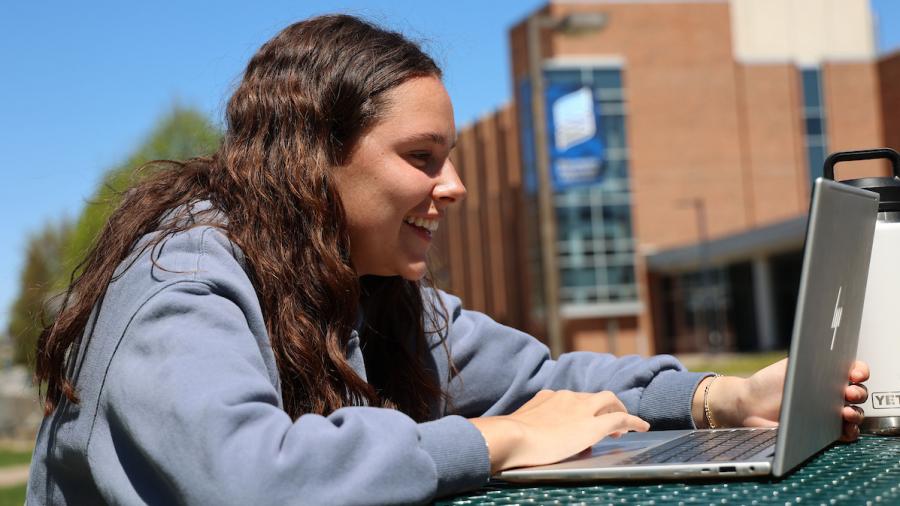 A student works at a lap top, sitting at a picnic table on campus.