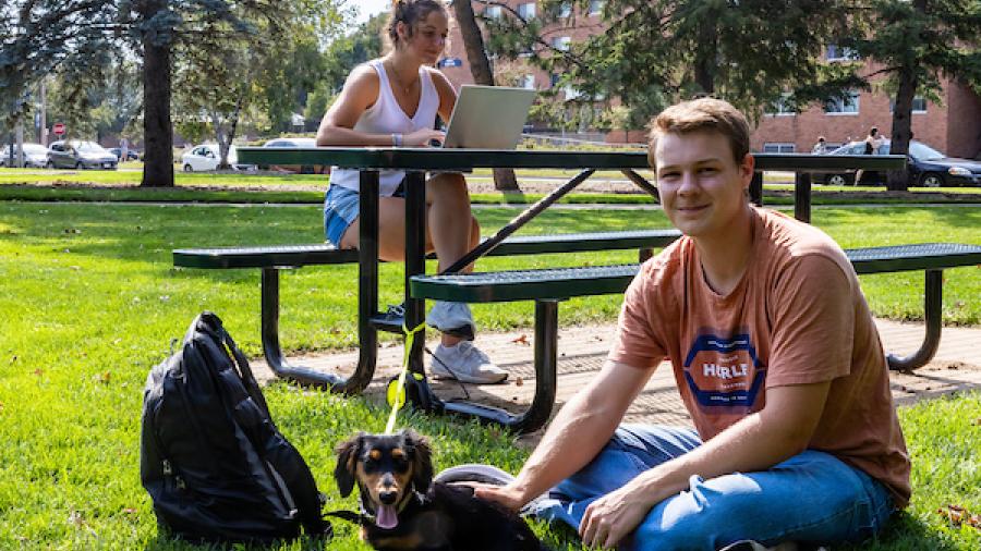 A student sits on the lawn with a small dog, while another works on a laptop at a picnic table.