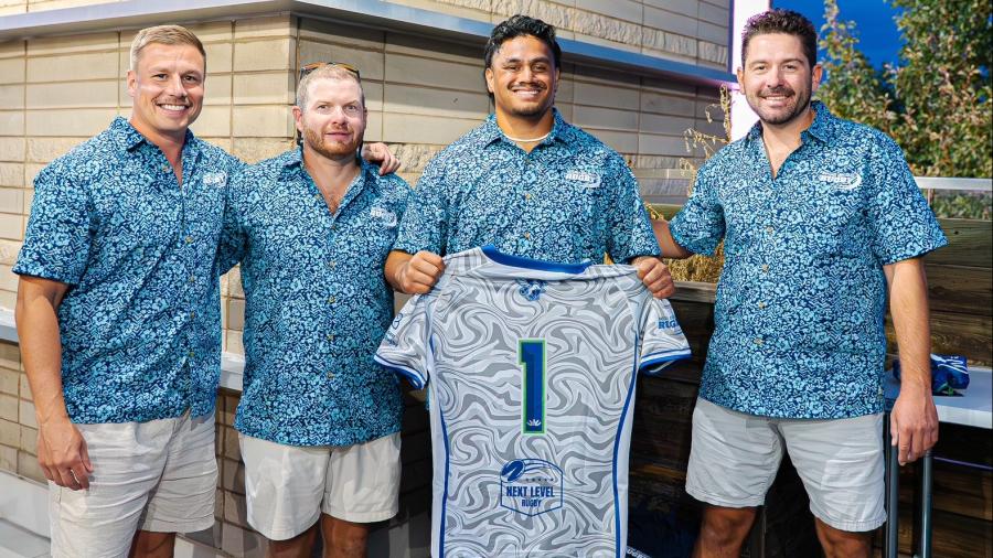 Four people standing together outdoors, all wearing matching blue patterned shirts and light-colored shorts. The two people in the center are holding up a gray and blue rugby jersey with the number one on it.