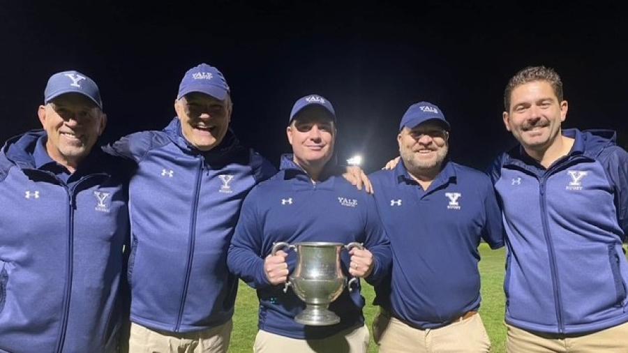 A group of five people standing on a sports field at night, all wearing matching navy jackets and khaki pants. The person in the center is holding a large silver trophy while the others stand close with arms around one another.