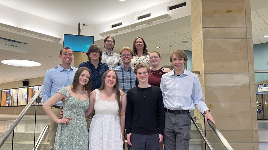 A group of people poses together on an indoor staircase. They are dressed in semi-formal attire and arranged in two rows with a brightly lit lobby area in the background.