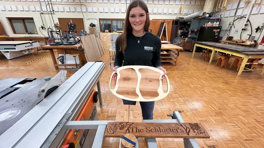 A student stands in a woodshop holding a wooden serving tray with multiple compartments. In front of them on a table is another finished wood piece engraved with decorative text. Large woodworking machines and worktables fill the background.
