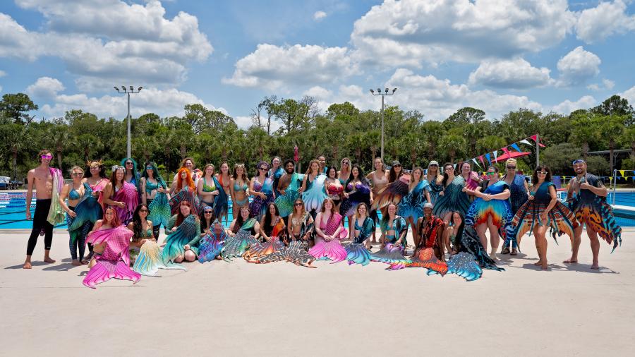 A large group of people wearing colorful mermaid tails posing together on the pool deck under a blue sky with scattered clouds.