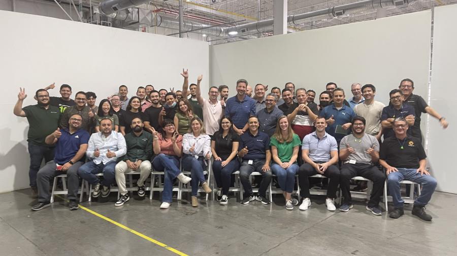 A large group of people poses together inside a spacious industrial warehouse. Some are seated in front while others stand behind them, many smiling and gesturing enthusiastically. The background shows high ceilings, exposed pipes, and bright overhead lighting.