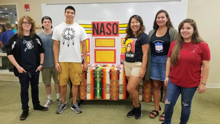 Several students stand in front of a colorful display table for the Native American Student Organization, featuring a large NASO sign and informational materials. If you'd like, I can also format them for a website, social media, or an accessibility report.