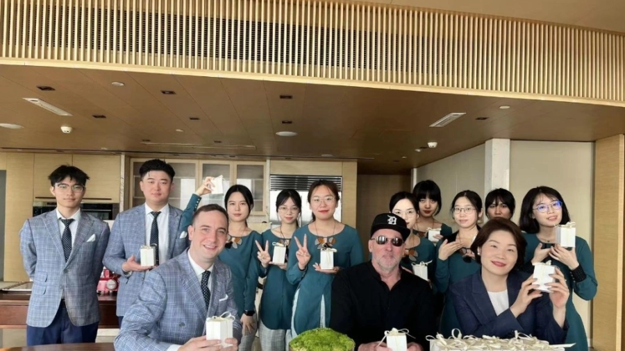 A large group of people stands together in a modern kitchen-like space with wood accents and a long wooden table in front of them. Many individuals are holding small, neatly wrapped gift boxes with ribbons. Some people in the back row are making peace signs with their hands. Two people sit at the table in the foreground, each holding a similar gift box, with a decorative bowl of greenery placed between them. More identical gift boxes are lined up on the right side of the table. The setting appears bright an