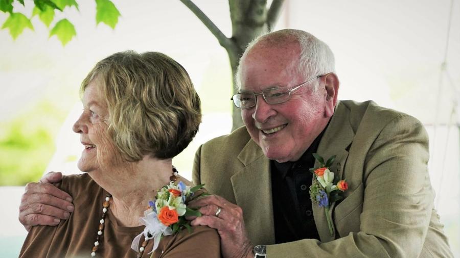 Two older adults seated outdoors under a tent, both wearing boutonnieres; one person places a hand on the other’s shoulder.