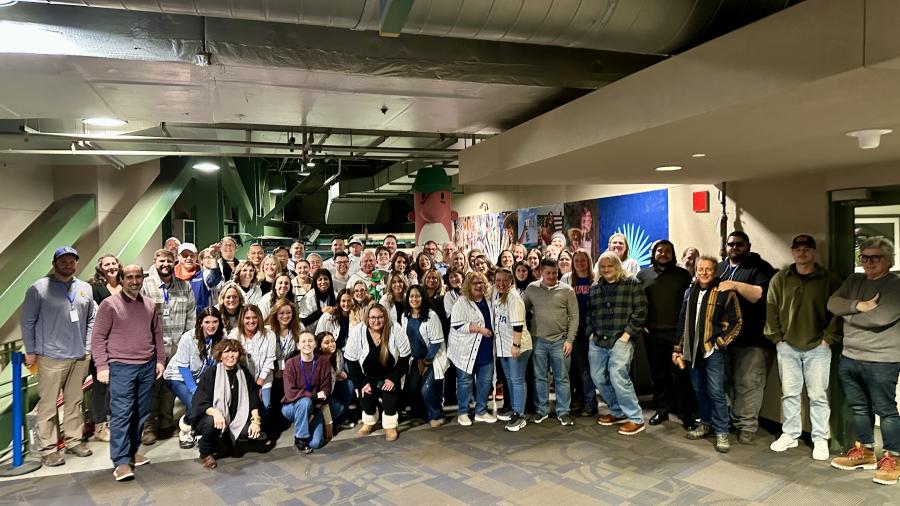 A large group of people poses together in an indoor venue with industrial-style ceilings and overhead ductwork. The group is arranged in several rows, with murals and signage on the wall behind them.