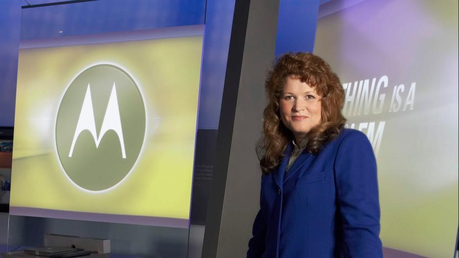 A person dressed in a blue suit stands at a modern presentation podium inside a studio-like setting. Behind them is a large screen displaying a Motorola logo and part of a slogan. The environment includes sleek, angular surfaces and professional lighting.