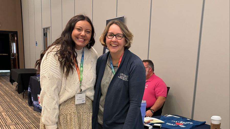 Two individuals stand together in a conference setting next to a University of Wisconsin–Stout display table. The table is covered with UW–Stout materials, brochures, and branded items. The individuals wear conference badges, and one person behind the table is seated. The room has patterned carpet, neutral-colored walls, and additional tables lining the hallway.