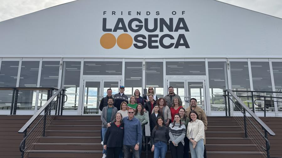 A group of people standing on outdoor wooden steps in front of a building with a large sign reading “Friends of Laguna Seca.”