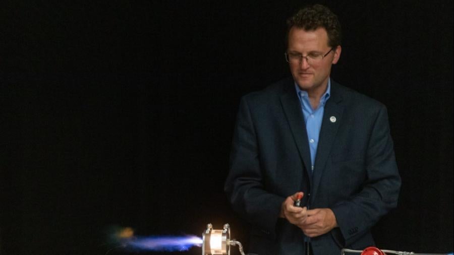 A person stands behind a table demonstrating a small rocket propulsion device, producing a bright blue flame, with control equipment and a gas tank arranged on the table against a dark background.