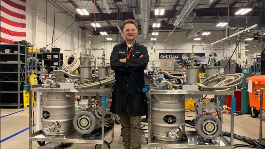 A person stands with arms crossed between two stainless steel vacuum cart ground support equipment units on wheeled frames inside an industrial aerospace facility. Each cart has hoses, valves and gauges, with high ceilings, ductwork and an American flag visible in the background.