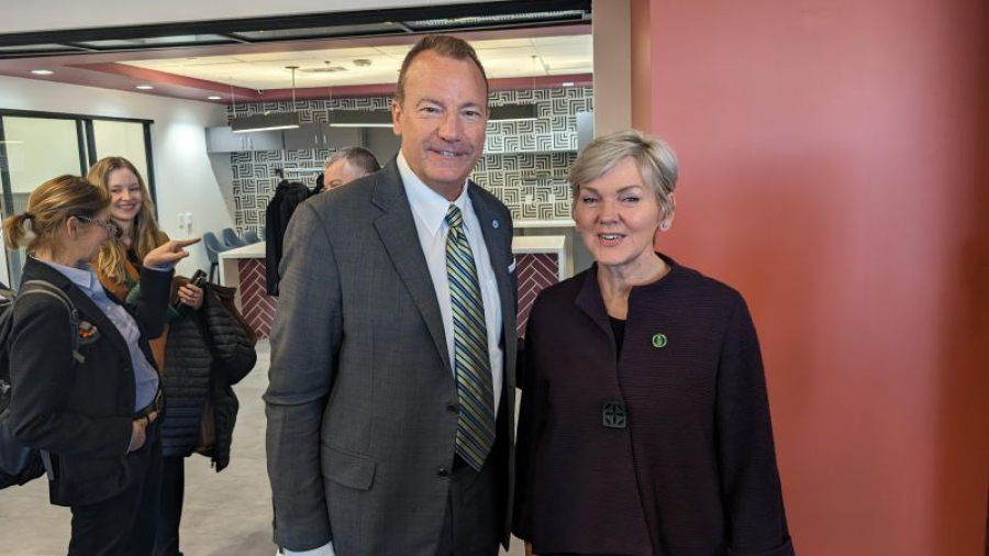 Two adults in business attire stand together in a modern lobby, facing the camera, with patterned wall décor and several people conversing in the background.