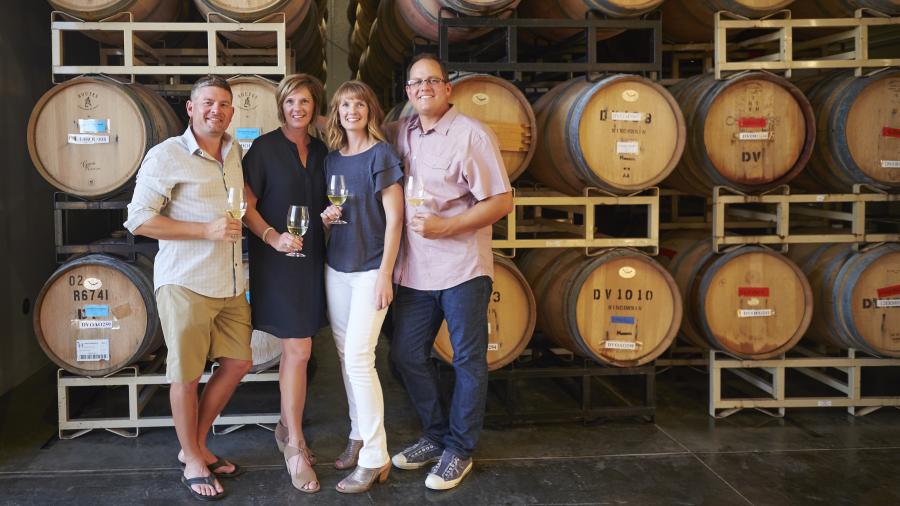 Four people standing in a winery in front of stacked wooden barrels, each holding a glass of white wine.