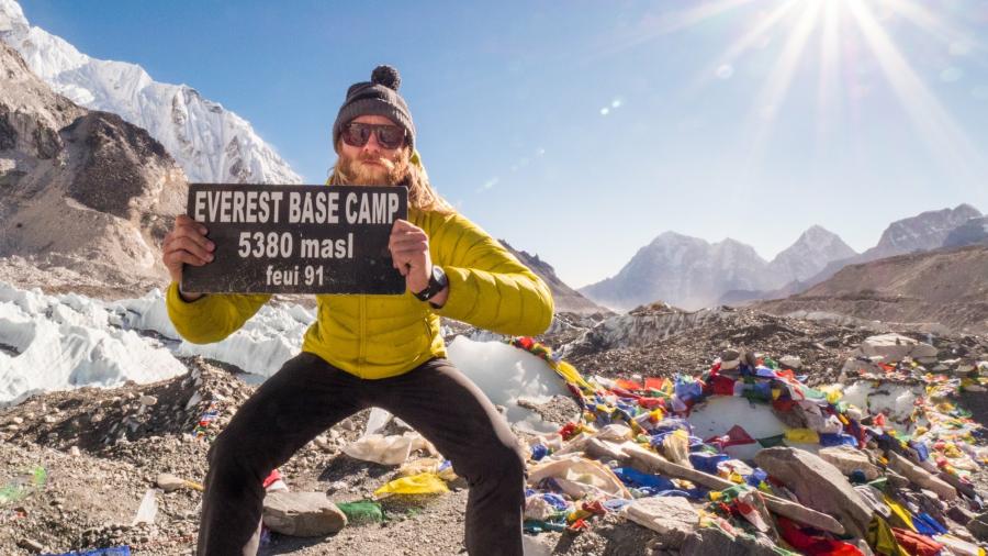 Person in a yellow jacket holds a sign reading “Everest Base Camp, 5380 masl” with snow-covered mountains and prayer flags in the background.
