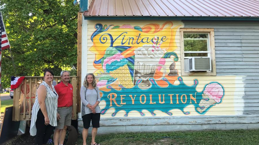 Three people stand beside a colorful “Vintage Revolution” mural painted on the exterior wall of a small building.