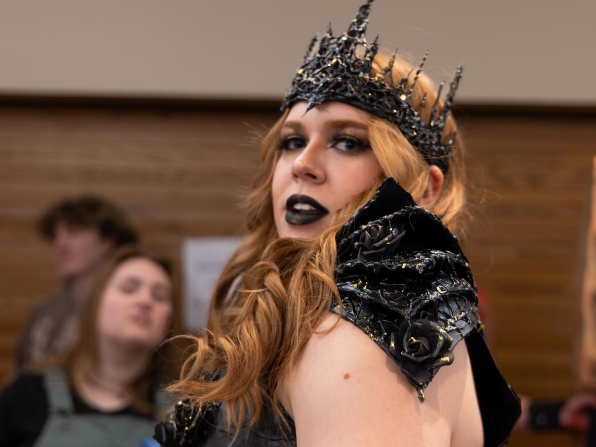 Female student in her project outfit turns and looks over her shoulder at the camera. She has a black spikey crown, with spike black pieces covering her shoulders.