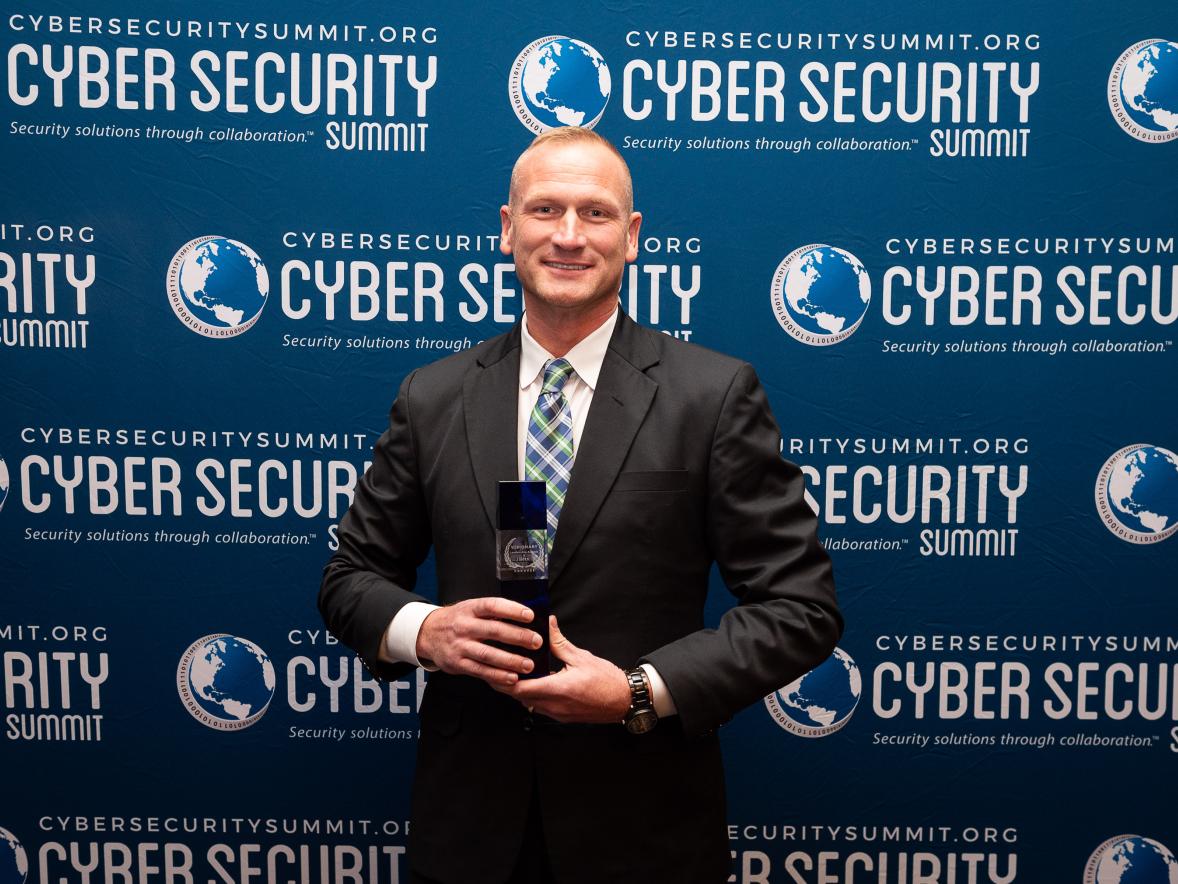 A person in a suit stands in front of a Cyber Security Summit step‑and‑repeat backdrop while holding an award.