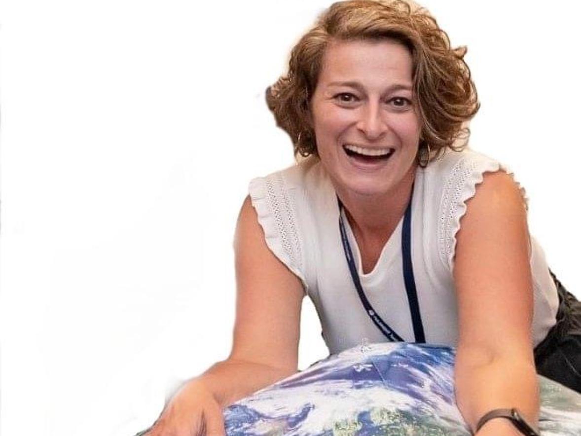 A person is leaning forward over a large inflatable globe, resting their hands on it while smiling toward the camera. They are wearing a sleeveless white top and a lanyard, with a watch on one wrist. The background is plain white.