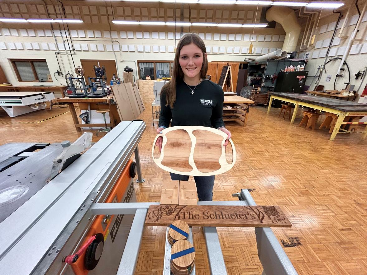 A student stands in a woodshop holding a wooden serving tray with multiple compartments. In front of them on a table is another finished wood piece engraved with decorative text. Large woodworking machines and worktables fill the background.