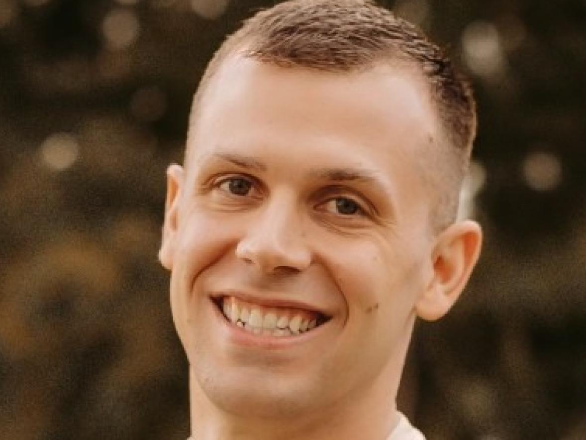 A person stands outdoors wearing a light-colored T-shirt. Trees and greenery form a soft, blurred background behind them.