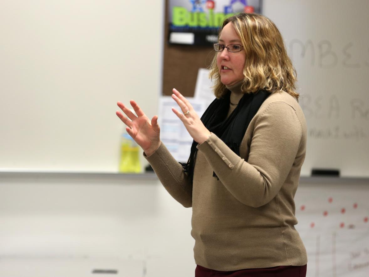 A person stands at the front of a classroom with hands raised mid-gesture. The individual is wearing a light-colored long-sleeve top and a dark scarf. Behind them is a whiteboard with faint writing and a bulletin board displaying classroom materials.
