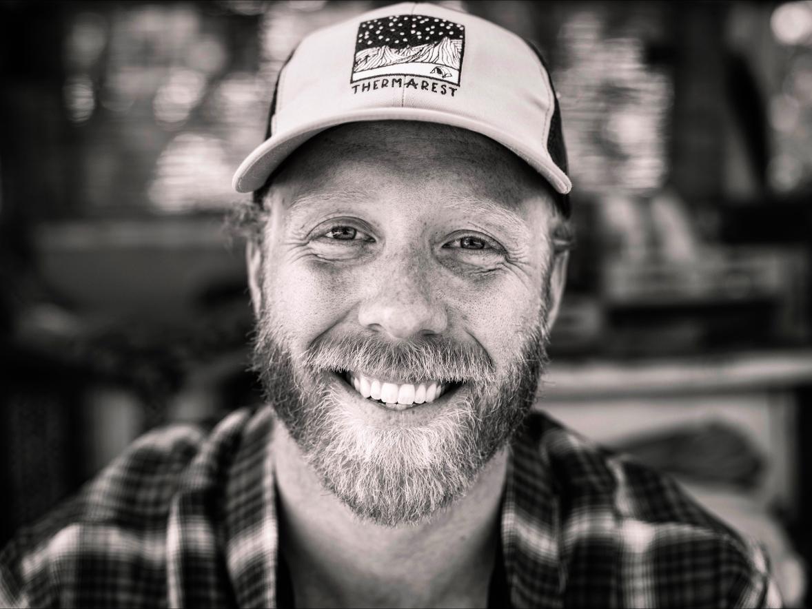 Close-up black-and-white portrait of a person with a beard wearing a cap, smiling toward the camera.