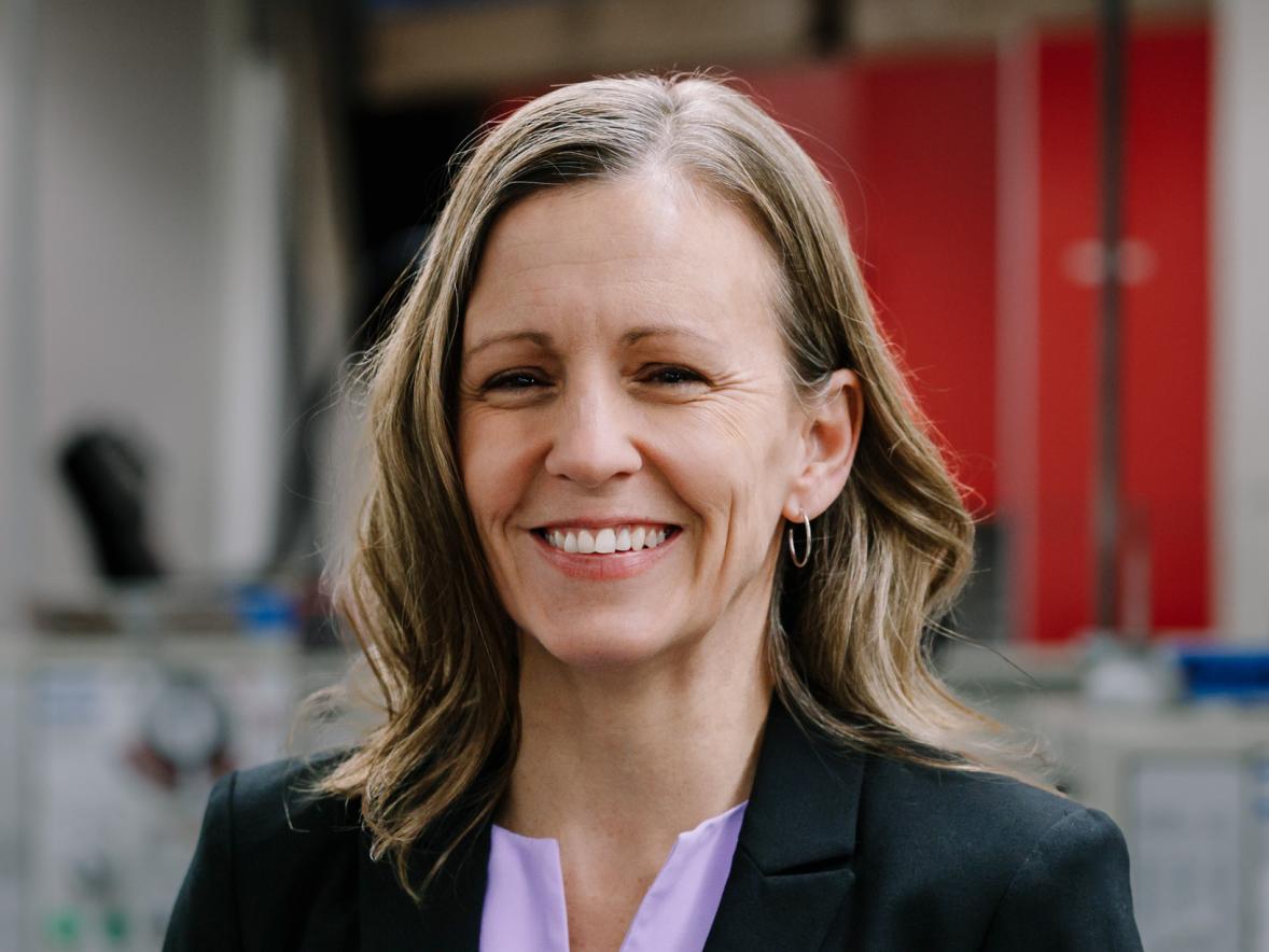 Person in a black blazer and light purple blouse, smiling at the camera, standing in an indoor lab or workspace with blurred equipment and red storage cabinets in the background.