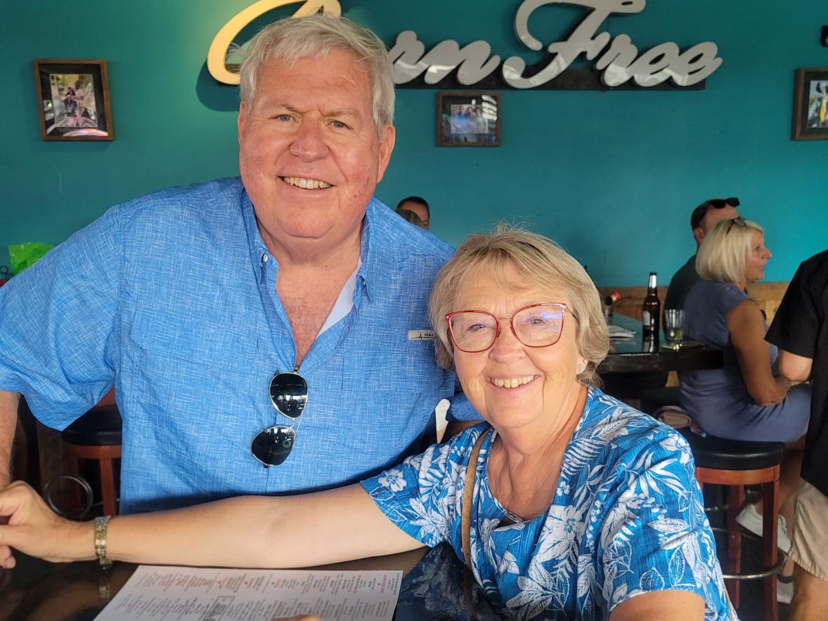 Two older adults seated at a restaurant table, smiling at the camera, with a turquoise wall and “Green Tree” sign behind them.