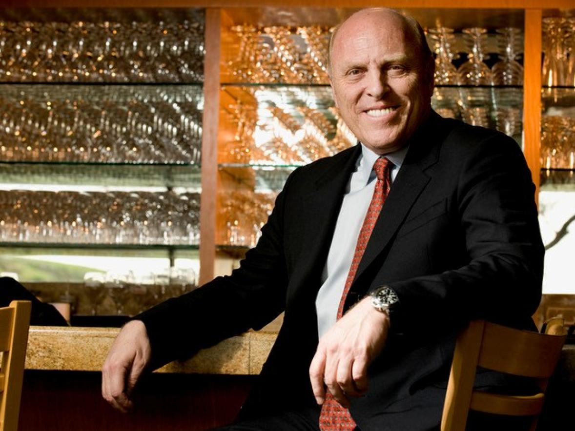 A person in a dark suit and red tie sits at a restaurant bar, with shelves of glassware displayed behind.