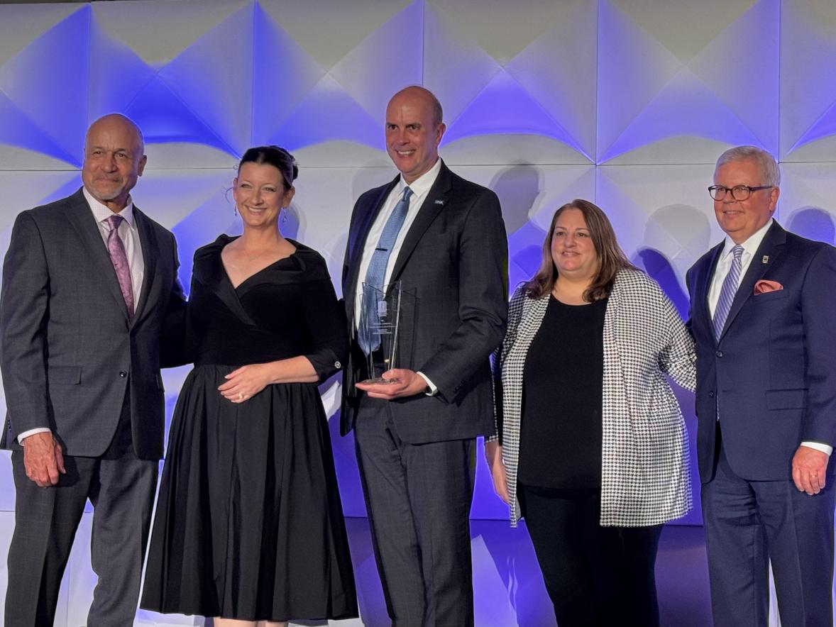 Five people pose onstage at an awards ceremony. One person in a dark suit holds a glass award while standing in front of a geometric wall illuminated with blue and purple lighting.